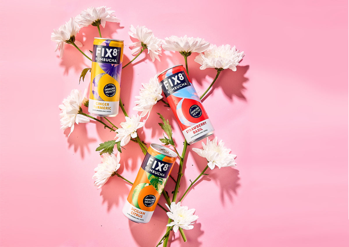 Three Fix8 kombucha cans laid flat among white chrysanthemums on a pink background